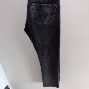 Levi's 501 svarta jeans - Svarta Levi's 501 jeans med klassisk femficksdesign och raka ben. Jeansen har en snygg tvättad finish, silverfärgad knapp och nitar samt den ikoniska läderpatchen bak i midjan. Perfekta för en avslappnad och stilren look.