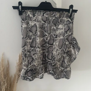 Grå kortkjol med ormmönster från Boohoo - Säljer en snygg kortkjol från Boohoo med grått och svart ormmönster. Kjolen har volangkant nertill och omlott-look på framsidan. Perfekt för dig som vill sticka ut med en trendig och edgy stil.