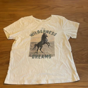 Vit t-shirt med hästmotiv från KappAhl - Vit t-shirt från KappAhl med coolt tryck av en stegrande häst och texten 'True Wilderness Dreams' på bröstet. Rund hals och korta ärmar, tillverkad i mjuk bomull. Perfekt för dig som gillar djur och naturvibbar.