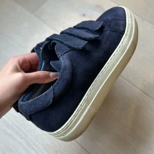Mörkblå sneakers från J.Lindeberg - Snygga mörkblå sneakers från J.Lindeberg i mocka och tre kardborreband. Populära skor. Skorna har en klassisk rund tå och är tillverkade i Portugal. Perfekta för dig som gillar stilrena och bekväma skor med en twist. Saknar sulor , ta ett par gamla sulor eller köpa nya