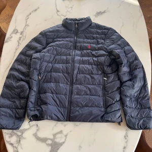 Mörkblå dunjacka Polo Ralph Lauren M - Snygg mörkblå dunjacka från Polo Ralph Lauren med quiltad design och dragkedja framtill. Jackan har ståkrage, två fickor med dragkedja och den klassiska röda loggan broderad på bröstet. Perfekt för kyliga dagar och enkel att matcha med allt. Skriv vid minsta funderingen!