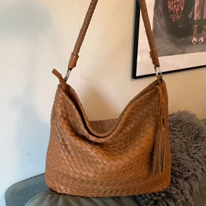 Brun axelväska i flätat skinn - Snygg brun axelväska från  Collezione Alessandro i flätat skinn. Väskan har en bred axelrem, tofsdetalj och rymligt innerfack med dragkedja. Perfekt för dig som gillar en stilren men ändå trendig look.