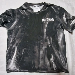 Svart och vit t-shirt från Moschino - Cool svart t-shirt från Moschino med unikt vitt mönster som ser ut som penseldrag. Kortärmad modell med rund hals och Moschino-logga tryckt på bröstet. Perfekt statement-plagg för dig som gillar streetstyle. Äkta