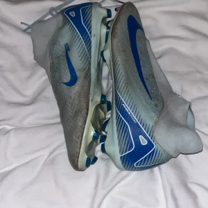 Nike mercurial pro, ljusblå fotbollsskor Z - Säljer ett par Nike fotbollsskor i grått med blå detaljer och stora blå swoosh-loggor. Skorna har en strumpliknande passform och mönstrad ovandel i syntetmaterial. Sulan har fasta dobbar för spel på gräs. Perfekta för dig som vill sticka ut på planen.