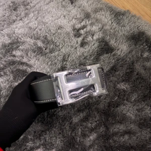 Lyxigt Svart H bälte hög kvalitet  - Snyggt svart bälte från H med klassiskt H-formad spänne i silverfärg. Bältet är gjort i präglat skinn med tydliga sömmar längs kanterna. Perfekt accessoar för att lyfta din outfit med en lyxig touch.