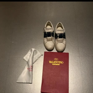 Valentino Garavani Open sneakers, vit/blå - Säljer ett par snygga Valentino Garavani Open sneakers i vitt skinn med bred blå rem över vristen och ikoniska nitar på hälen. Klassisk rund tå, platt sula och snörning. Skorna kommer med originalkartong och dustbag. Pris kan diskuteras. Hör av er vid frågor!