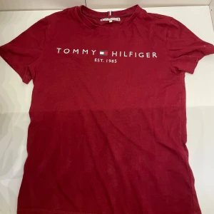 Vinröd t-shirt från Tommy Hilfiger 152cm - Snygg vinröd t-shirt från Tommy Hilfiger i storlek 152 cm. Tröjan har den klassisk loggan och text framtill. T-shirten har rund halsringning och korta ärmar. Tillverkad i mjuk bomull som är skön att bära. Perfekt för en stilren och avslappnad look. Mvh Hannes!
