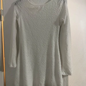 Vit spetsklänning med blommönster - Superfin vit kortklänning med lång ärm och blommigt spetsmönster. Klänningen har rund halsringning, är fodrad och har transparenta ärmar. Perfekt för dig som gillar romantisk och feminin stil.