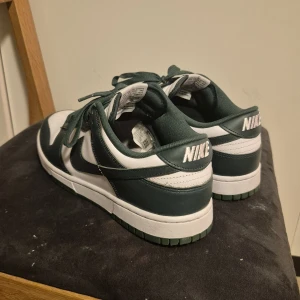 Nike Dunk Low gröna och vita sneakers - Säljer ett par Nike Dunk Low sneakers i grönt och vitt skinn med klassisk Nike-logga på sidan och hälen. Skorna har grön yttersula, snörning och perforerad tå för extra ventilation. Perfekt för dig som gillar streetwear och vill sticka ut med snygga färger. Storlek 44, nyskick använd typ 3 ggr