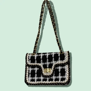 Rutig axelväska med kedjerem - Snygg axelväska i svart och vitt med rutigt mönster och dekorativ flätad kant. Väskan har en guldfärgad kedjerem och ett vridlås framtill. Perfekt för dig som vill ha en trendig och stilren accessoar med lite extra edge.