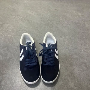 Mörkblå sneakers från Hummel - Snygga mörkblå sneakers från Hummel med vita detaljer och klassisk logga på sidorna. Skorna har snörning, vit sula och är tillverkade i textil och syntet. Perfekta för dig som gillar sportig och stilren look.