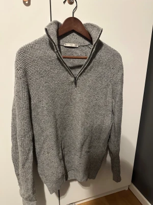 Grå stickad half zip-tröja  - Säljer en grå stickad tröja från Mango i storlek S. Tröjan har en grovstickad struktur, hög krage och half zip-dragkedja framtill. Långärmad och med ribbade muddar vid ärmslut och nederkant. Perfekt för kyliga dagar.