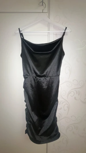 Svart satinklänning med smala axelband - Svart kort satinklänning med smala justerbara axelband och lätt draperad urringning. Klänningen har en glansig finish och figurnära passform, perfekt för dig som gillar en stilren och trendig look. Bra material har inte använt den jag köpte den för några dagar sen och det va inte min storlek. Den är tvättad och ren.