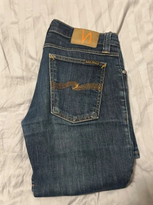 nudie jeans mörkblåa -   Snygga mörkblå jeans från Nudie Jeans, modell Tight Long John. De har klassisk femficksdesign, kontrastsömmar och en smal passform med stretch för extra komfort. Materialet är 98% bomull och 2% lycra. Perfekt för dig som gillar en tight och modern look. Midja 36cm, längd 100cm. 