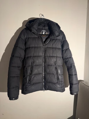 Svart pufferjacka från Save The Duck - Svart pufferjacka från Save The Duck med huva och två sidofickor med dragkedja. Jackan är quiltad och har syntetiskt Plumtech-foder som är animal free. Perfekt för kalla dagar och har en clean, modern look. Jackan är sparsamt använd och i ett bra skick. Hör av dig vid minsta fråga eller prisfråga!