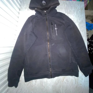 Svart Sail Racing hoodiejacka 150 - Svart hoodiejacka från Sail Racing med dragkedja framtill och bröstficka med zip. Jackan har huva med logga, långa ärmar och är tillverkad i en mjuk blandning av bomull och polyester. Perfekt för chill dagar och streetwear.