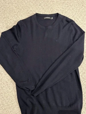 Svart merino tröja från J.Lindeberg - Svart stickad tröja från J.Lindeberg i storlek M. Klassisk rund hals och diskret logga på bröstet. Tröjan har långa ärmar och ribbade muddar vid ärmslut och nederkant. Perfekt för dig som gillar stilrena och enkla plagg.