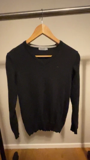 Svart stickad tröja från J.Lindeberg - Svart stickad tröja från J.Lindeberg i storlek S. Tröjan har v-ringad hals och långärmade ärmar, med en liten broderad logga på bröstet. Perfekt för dig som gillar stilrena och klassiska plagg med snygg passform.