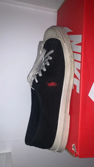 Svarta Polo Ralph Lauren sneakers - Svarta sneakers från Polo Ralph Lauren med vit platt sula och vit gummitå. Skon har vit snörning och röd Polo-logga broderad på sidan samt tryckt på innersulan. Ovandelen är i canvas och sulan i gummi, med rund tå och klassisk streetstil.