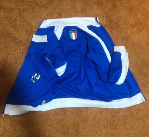 Blå Puma vindjacka Italien M - Blå vindjacka från Puma med Italiens landslagsmärke på bröstet och vita detaljer vid muddar och krage. Jackan har broderade guldstjärnor och dragkedja. Perfekt för dig som gillar fotboll och vill ha en sportig stil.