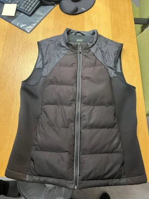Svart dunväst från Hugo Boss - Snygg svart dunväst från Hugo Boss med quiltad framsida och dragkedja. Västen har hög krage och insidan är dekorerad med Boss-logga och mönster. Materialet är lätt och stretchigt med fyllning av ankdun. Perfekt för lager-på-lager under kyliga dagar.
