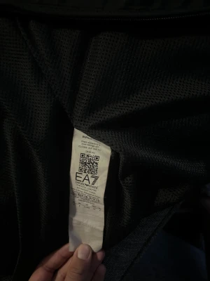 Svart vindjacka från EA7 Emporio Armani - Svart vindjacka från EA7 Emporio Armani med huva och meshfoder. Jackan har dragkedja framtill och EA7-logga på bröstet. Snygg och sportig design som passar perfekt för dig som gillar stilrena och funktionella plagg.