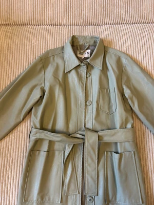 Grön overshirt jacka från NA-KD - Snygg ljusgrön overshirt jacka från NA-KD i syntetmaterial med skinnlook. Jackan har stora fickor fram, knappar hela vägen och ett matchande bälte i midjan. Klassisk krage och långärmad modell, perfekt för lager på lager. Stilren och trendig design. Nypris 600kr.