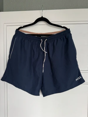 Marinblå shorts från BOSS med snörning - Snygga marinblå shorts från BOSS med vit snörning i midjan och logga broderad på benet. Modellen har elastisk midja, fickor på sidorna och en bakficka. Perfekta för chill dagar eller träning, tillverkade i lätt polyester som andas.