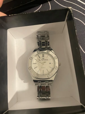 Emporio Armani silverfärgad klocka - Snygg klocka från Emporio Armani med silverfärgat armband i metall och oktagonformad boett. Urtavlan är ljus med subtilt mönster och har datumvisning. Klockan har en stilren design med markerade skruvar på boetten och klassisk länkarmband.