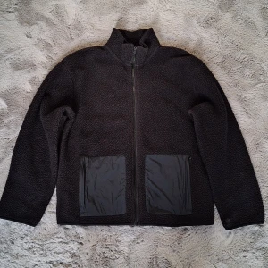 Pilejacka/teddyjacka i nyskick stl 158/164 - Svart teddyjacka från H&M med två stora fickor framtill i ett slätt material. Jackan har hel dragkedja och hög krage, perfekt för kyliga dagar. Materialet är mjukt och fluffigt, vilket ger en skön och avslappnad vibe. Passformen är normal och jackan är långärmad.