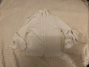 Vit ribbad träningsskofta med tumhål - Säljer en stilren vit ribbad träningskofta med dragkedja hela vägen och hög krage. Tröjan har långa ärmar med tumhål, vilket ger en sportig vibe. 