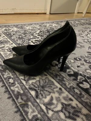 Svarta pumps från Graceland, storlek 37 - Snygga svarta pumps från Graceland i storlek 37. Skorna har spetsig tå, hög klack och är tillverkade i syntetmaterial med en slät finish. Sulan har ett diskret blommigt mönster för extra detalj. Perfekt för dig som gillar klassisk och stilren look. Klacken är 11cm