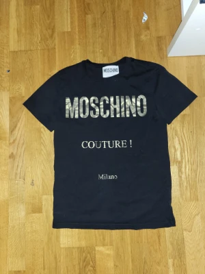 Svart Moschino Couture t-shirt - Svart t-shirt från Moschino med guldigt tryck på bröstet där det står 'MOSCHINO COUTURE! Milano'. Klassisk rund hals och korta ärmar. Tillverkad i 100% bomull för en skön och mjuk känsla. Perfekt för dig som gillar statement-plagg och exklusiv design.