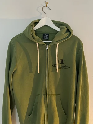 Grön zip hoodie Champion, medium - Grön zip-hoodie från Champion i storlek medium. Tröjan har huva med vita snören, broderad logga på ärmen och tryckt Champion Athletic Dept på bröstet. Mjuk bomullsmix, två fickor fram och ribbade muddar. Perfekt för chill eller streetwear.