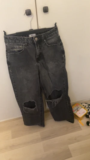 Högmidjade Svarta ripped jeans från Only - Säljer ett par svarta jeans från Only med slitna hål över knäna. Jeansen har klassisk femficksdesign, knapp och dragkedja framtill samt en tvättad, lite blekt look. Perfekta för dig som gillar en edgy och avslappnad stil.