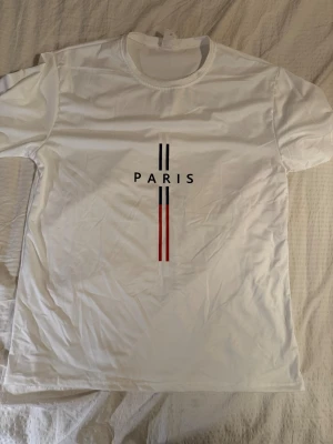 Vit t-shirt med Paris-tryck - Vit t-shirt med trycket 'PARIS' och två svarta samt två röda vertikala ränder på framsidan. Klassisk rund hals och korta ärmar. Tillverkad i mjuk bomull, perfekt för en clean och stilren look.