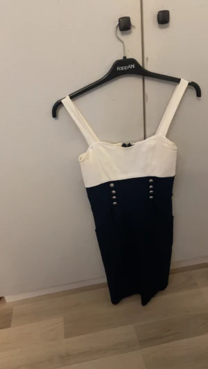 Marinblå och vit klänning med knappar - Snygg kort klänning med breda axelband. Överdelen är vit och nederdelen marinblå, dekorerad med åtta silvriga knappar framtill. Klänningen har en figurnära passform och dragkedja bak. Perfekt för dig som gillar stilren och klassisk look.