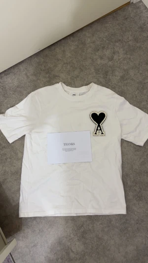 Vit t-shirt från Ami Paris med hjärtdetalj - Vit t-shirt från Ami Paris med stor svart hjärta och A-logga på bröstet. Klassisk passform, rund hals och korta ärmar. Tillverkad i mjuk bomull för en skön känsla. Perfekt statement-plagg för dig som gillar stilrena men unika detaljer. OBS! Den är använd men kommer med kvitto och påsen, inga skador