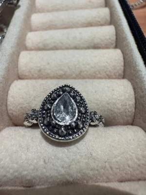 Silverring med droppformad sten - Unik silverring med en stor, droppformad klar sten i mitten och små dekorativa detaljer runt om. Ringen har en vintage vibe med pärlformade inslag på sidorna och runt stenen. Perfekt för dig som gillar statement-smycken med lite extra bling.