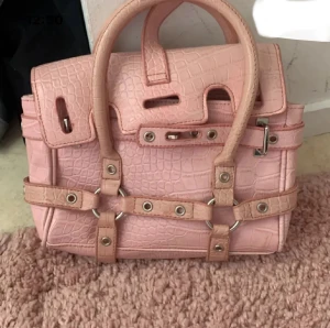 Rosa handväska med krokopräglat mönster - Säljer en snygg rosa handväska med krokopräglat mönster och coola metalldetaljer. Väskan har dubbla handtag, dekorativa remmar med nitar och spännen, samt en unik design som sticker ut. Perfekt för dig som vill ha en statement-väska i din samling.