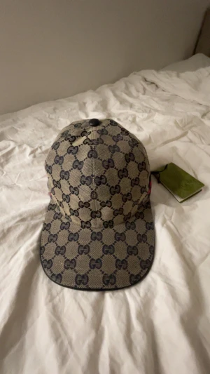 Blå gucci keps med blå röda stripez. keps med GG-mönster - Snygg beige keps från Gucci med klassiskt GG-mönster i mörkblått över hela kepsen. Kepsen har böjd skärm och detaljer i mörkblått längs kanten. Märkets logga syns tydligt och materialet är slitstark bomullscanvas. Perfekt för dig som gillar lyxiga accessoarer.