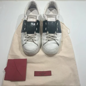 Valentino rockstud vita- svart glittriga sneakers.  - Valentino rockstud vita- svart glittriga sneakers. Snygga vita sneakers från Valentino Garavani med bred svart rem över vristen och coola nitar på hälen. Skorna är i skinn och har klassisk snörning. Kommer med originalpåse och äkthetskort. Perfekta för dig som vill ha en lyxig och trendig look. De är svartglittriga. De har blivit upprustade med läderfärg för att få sin svarta look. Man kan se detta om man tar bort snörningen men det är inte synligt från utsidan. Storlek 37