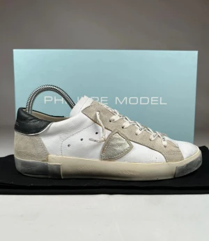 Philippe Model vita sneakers med silverdetalj - Snygga sneakers från Philippe Model i vitt skinn med beige mockadetaljer och svart häl. Skorna har en silverfärgad sköld på sidan och coola snören med text. Platt sula och rund tå ger en klassisk streetstil. Perfekta för dig som gillar exklusiva sneakers med unik design. Skoboxen och dustbagen ingår.