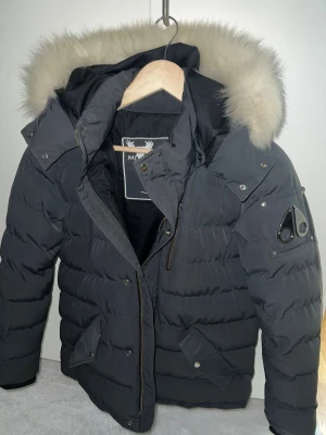 Grå Moose Knuckles dunjacka med päls (Limited ed)  - Snygg svart dunjacka från Moose Knuckles med fluffig ljus päls på huvan. Jackan har quiltad design, dragkedja och knappar framtill, samt två fickor med lock. Märkets ikoniska logga sitter på ärmen. Perfekt för kalla dagar. Jackan har använts få gånger, Storleken är barn storlek 164-170 ungefär, XS passad perfekt om man är lite smalare i kroppen, den är grå och unik.