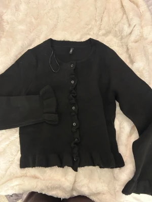 Svart kofta med volang från ONLY - Svart kofta från ONLY i stickat material, med volangdetaljer längs knappraden och vid ärmsluten. Rund halsringning och långa ärmar. Knappar framtill och ribbad kant nedtill. Perfekt för dig som gillar en söt och chill stil.