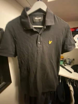 Svart pikétröja från Lyle & Scott - Svart pikétröja från Lyle & Scott med klassisk krage och två knappar framtill. Tröjan har det ikoniska gula fågelbroderiet på bröstet och är tillverkad i 100% bomull för skön komfort. Perfekt för en clean och stilren look.
