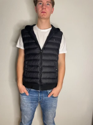 Svart dunväst med huva från Polo Ralph Lauren - Snygg svart dunväst från Polo Ralph Lauren med huva och dragkedja framtill. Västen har quiltad framsida, två fickor med knappstängning och diskret logga på bröstet. Baksidan är slät och i mjukt material. Perfekt för lager-på-lager under höst och vinter.