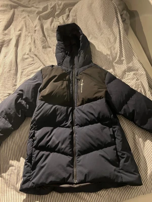 Mörkblå pufferjacka Sail Racing - Mörkblå pufferjacka från Sail Racing med svarta detaljer över axlar och bröst. Jackan har huva med logga, hel dragkedja framtill och två bröstfickor med dragkedja. Denna jacka är perfekt för kalla dagar och har en sportig look.