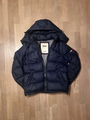 Marinblå pufferjacka från Tommy Hilfiger - Snygg marinblå pufferjacka från Tommy Hilfiger Denim med huva och dragkedja. Perfekt för kalla dagar och riktigt skön att bära ❄️. Personen på bilden är 183.   Skick 9/10 ✅ Nypris cirka 2500 Skickar inom 48 timmar om inte samma dag 🚚