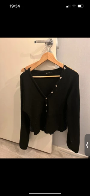 Svart kofta med knappar från Gina Tricot - Svart kofta från Gina Tricot i storlek M. Modellen har v-ringning och cool knappdetalj längs ena axeln och framtill. Långärmad och i mjukt stickat material, perfekt för lager-på-lager och snygg till både jeans och kjol.